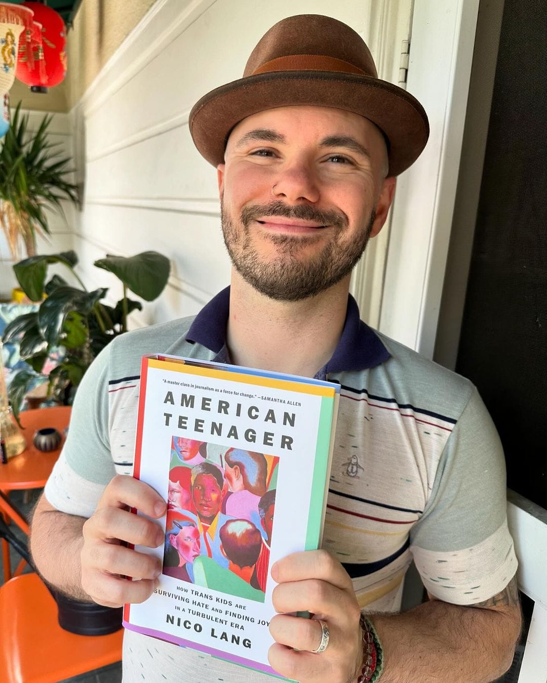 Nico Lang, author of American Teenager, is holding their book in their hands, smiling and looking at the camera. They are wearing a brown brimmed hat and the cover of the book is a multicolor portrait of multiple individuals. 