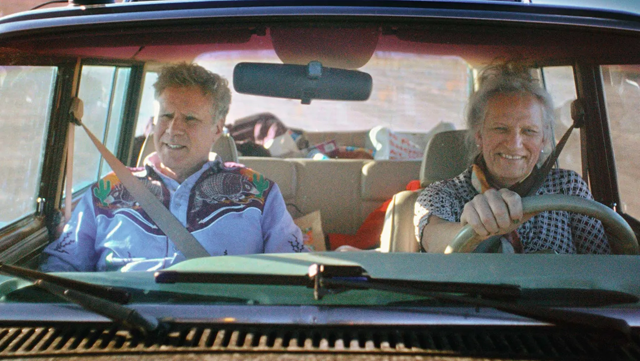 Will Ferrel and friend Harper in a car. Will is wearing a light blue button-up with embriodery while Harper is wearing a checkered short sleeve and scarf, 